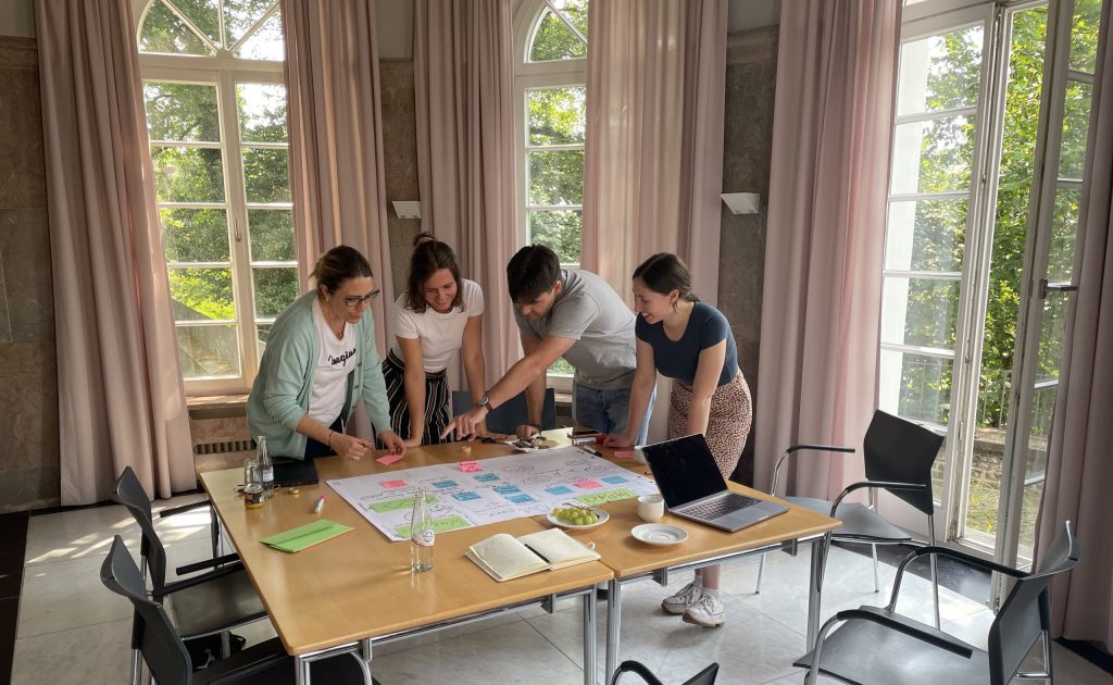 Vier Personen stehen vor einem Tisch und arbeiten zusammen an einem Flipchart